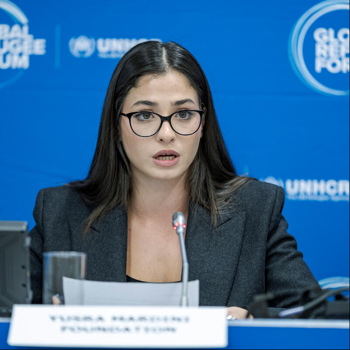 Una mujer joven, que usa anteojos, pronuncia unas palabras frente a un micrófono; detrás suyo, un fondo azul con el logotipo del Foro Mundial sobre los Refugiados.