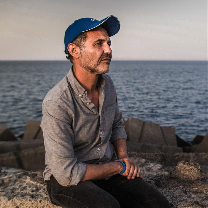 Sentado sobre unas rocas a la orilla del mar, un hombre que lleva puesta una gorra color azul mira al horizonte