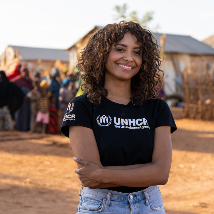 Una mujer afrodescendiente sonríe a la cámara con los brazos cruzados sobre el abdomen; detrás suyo, un grupo de personas en un campamento de refugiados