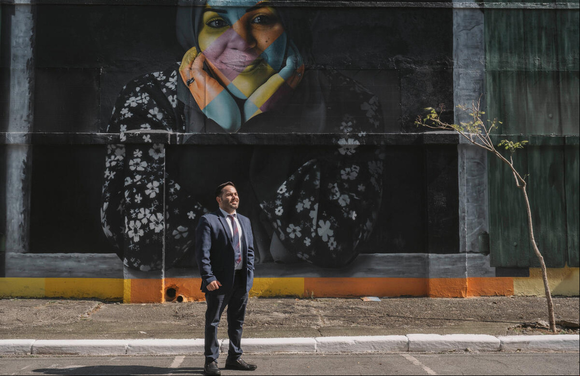 Un hombre vestido con traje y corbata de pie en una calle frente a un muro negro con el rostro de una mujer en colores