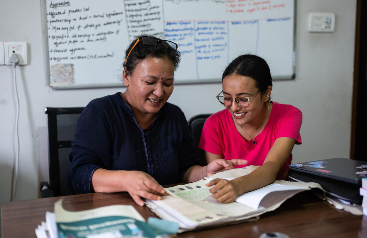 Dos mujeres nepalíes, sentadas frente a un escritorio, revisan un documento; detrás suyo, un pintarrón lleno de texto
