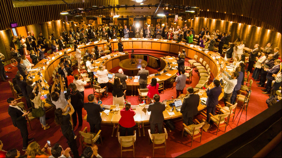 Panoraminc view of the conference room while delegates stand and clap their hands.
