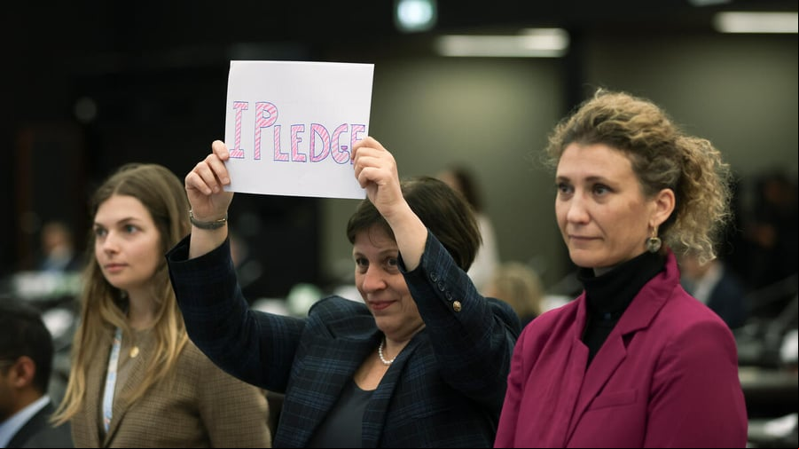 Tres mujeres forman una hilera. Las que están a los extremos son rubias; la que está en el centro, de cabello corto y oscuro, sostiene frente a sí un cartel que dice "Me comprometo".