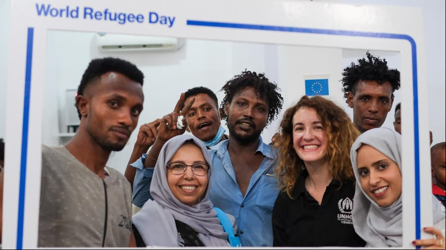 Un grupo de personas sonríe a través de un marco que simula una publicación en redes sociales y que en los márgenes dice "Día Mundial del Refugiado, ACNUR, #ConLosRefugiados"