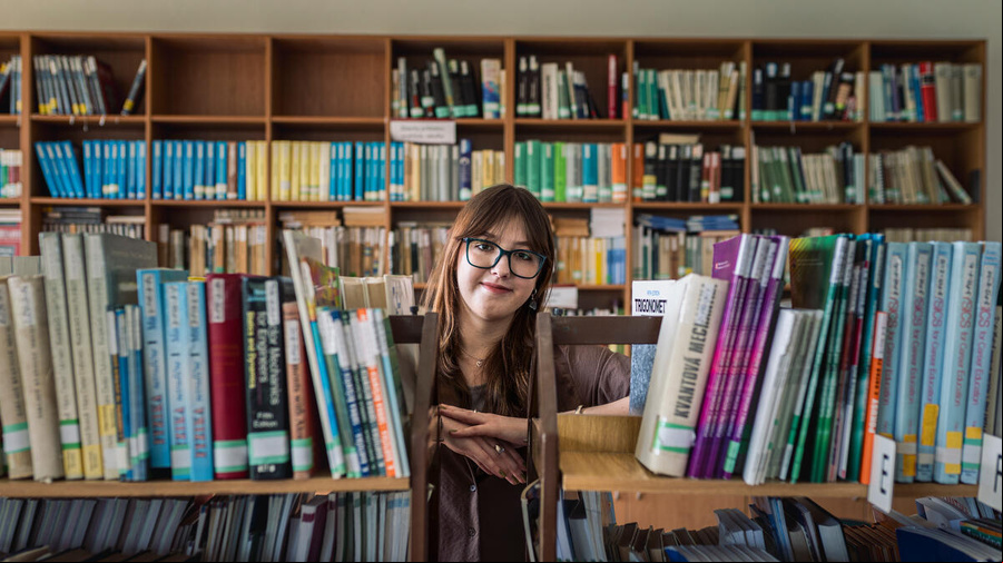 En una biblioteca repleta de libros, detrás de un par de estantes, una joven que usa anteojos y lleva el cabello suelto sonríe a la cámara.