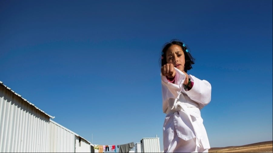 En un campamento de refugiados, una niña que lleva puesto un uniforme blanco de karateka hace un movimiento de karate frente a la cámara
